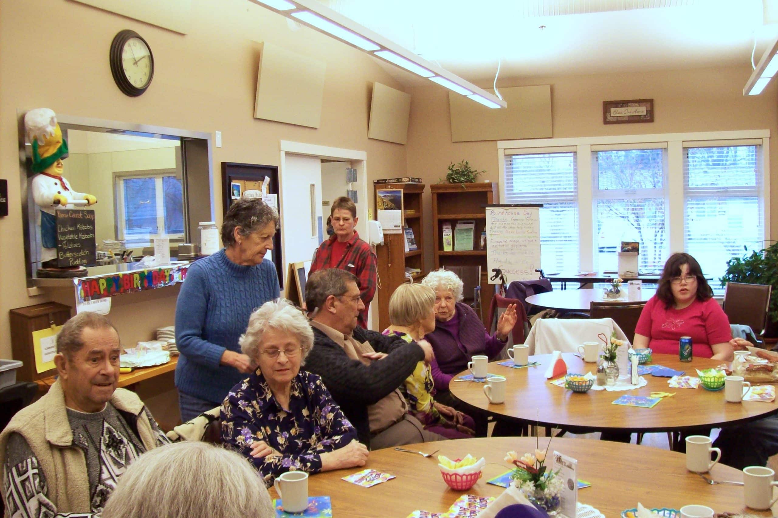 Seniors sit at tables drinking coffee and chatting in the community hall.