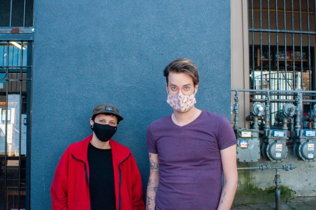 Local performing artists Jana Crook and Chris Thompson stand in front of a blue building with face masks on.