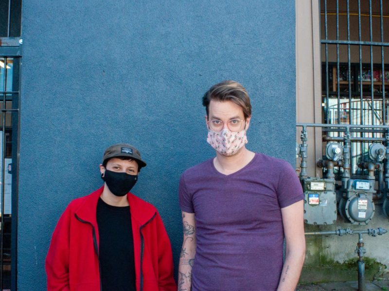 Local musicians Jana Crook and Chris Thompson stand in front of a blue building with face masks on.