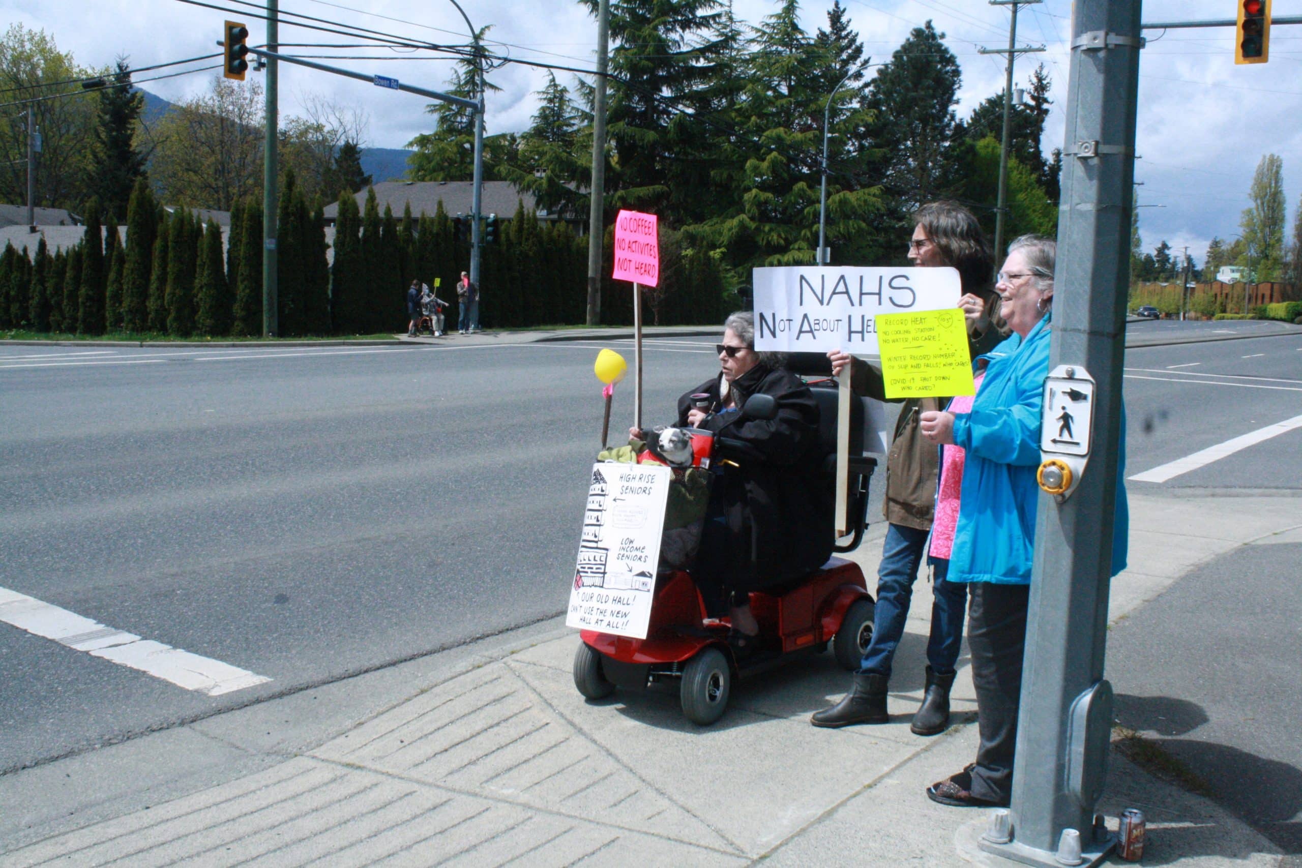 Three older women stand at the corner of Buttertubs Drive holding protest signs that read "No coffee, no activities, not heard."