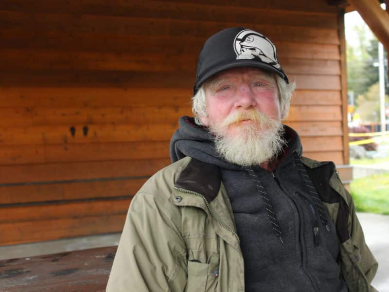 A portrait photo of Randy Thorensen, a white man with white hair and a beard. He is wearing a raincoat over a black jacket, with a black baseball cap on.