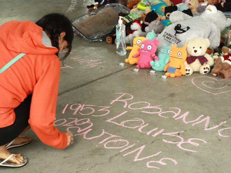 A person wearing orange writes a message in chalk near the donations of teddy bears that reads "Rosanna Louise Antoine 1953.
