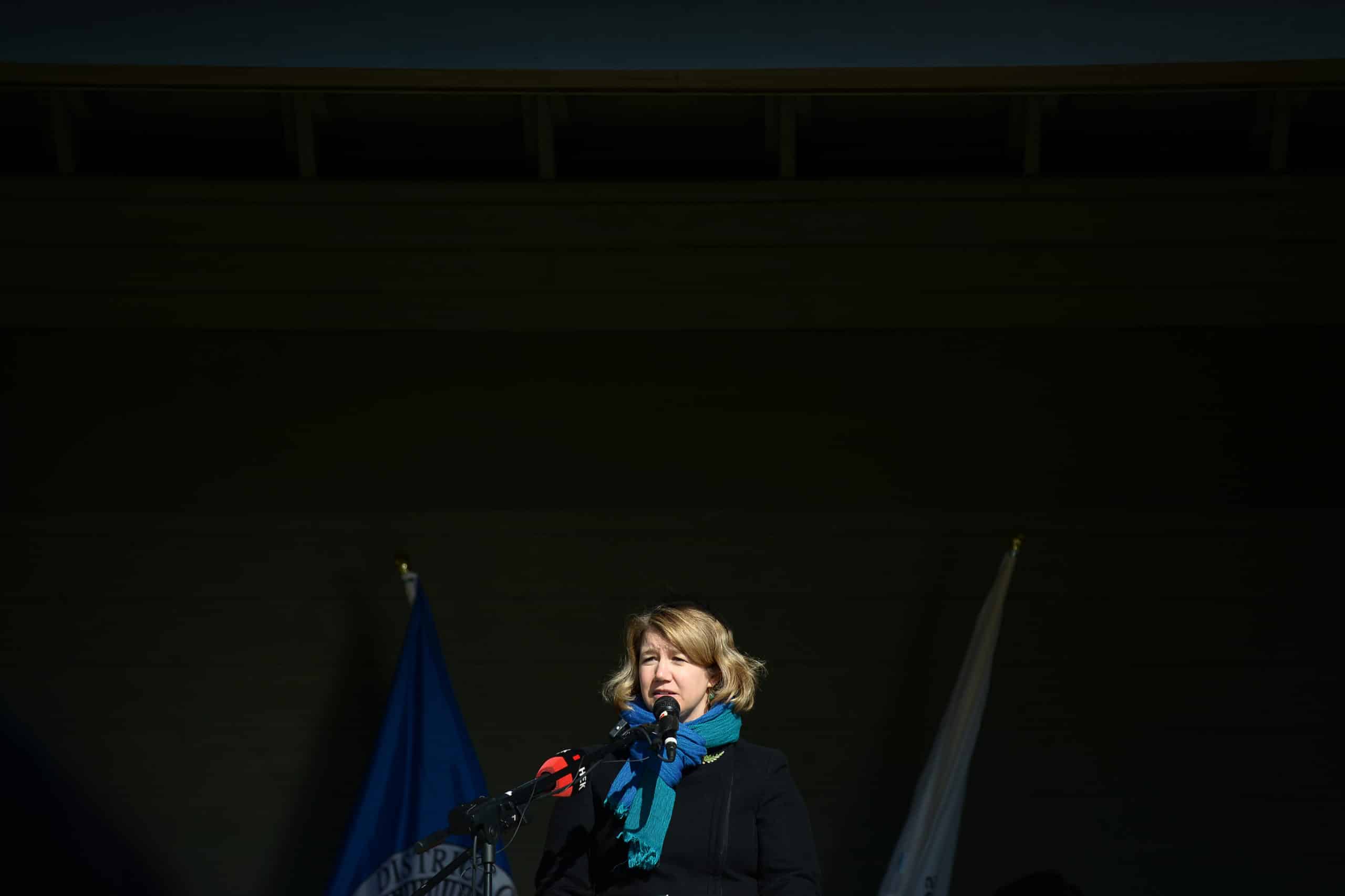Habitat Acquisition Trust executive director Katie Blake speaks on stage at the announcement event with two microphones in front of her. The background is nearly black and Blake's bright blue scarf stands out.