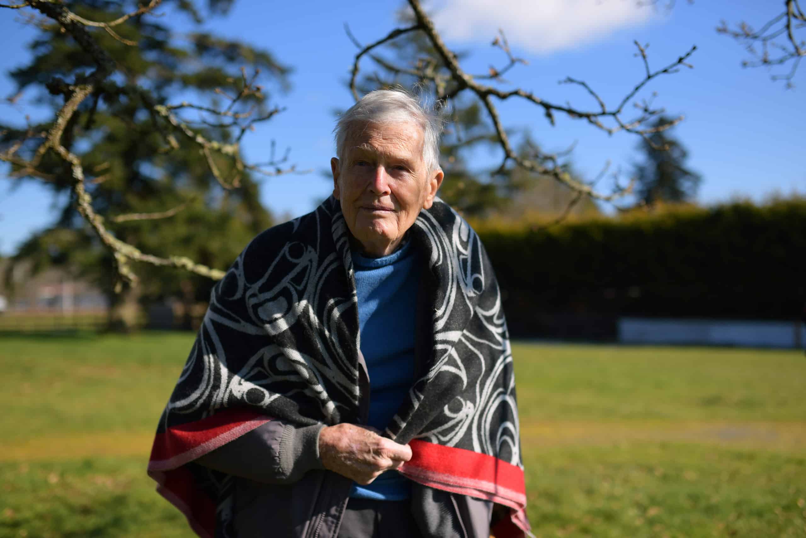Lifelong Metchosin resident Chris Pratt stands in front of trees and a grassy lawn. He is wrapped in a blanket with Coast Salish design.