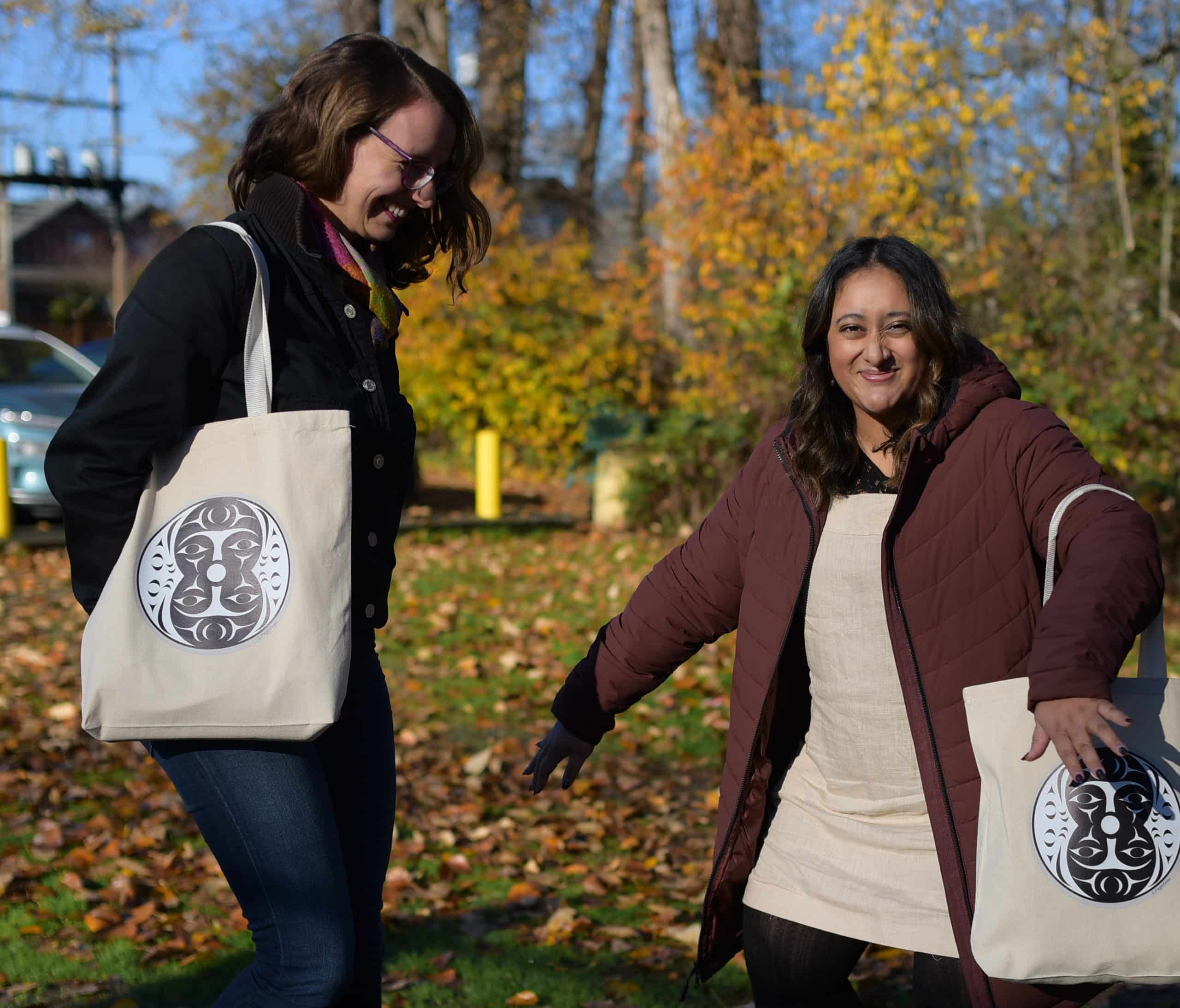 Jacqueline Ronson (left) and Shalu Mehta (right) laugh and dance in a silly moment. The two are carrying The Discourse tote bags and are outdoors, with trees in the background.