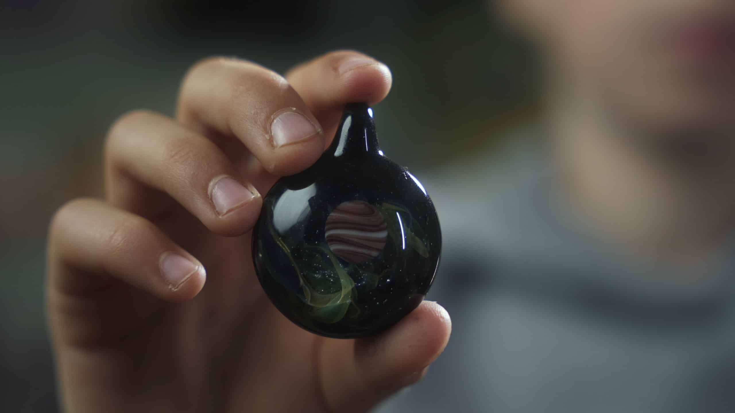 A had in the foreground holds an object that looks like a round glass bead with a hole in the middle. It is a still from the film being produced for Q'shintul/Mill Bay Nature School.