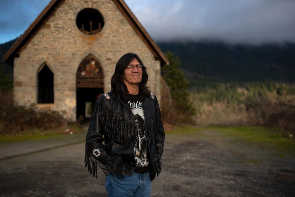 Harold Joe stands in front of an abandoned building in the foreground, with Mt. Tzouhalem seen in the background.