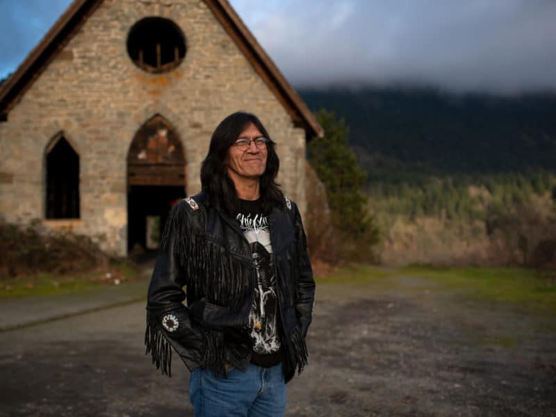 Harold Joe stands in front of an abandoned building in the foreground, with Mt. Tzouhalem seen in the background.