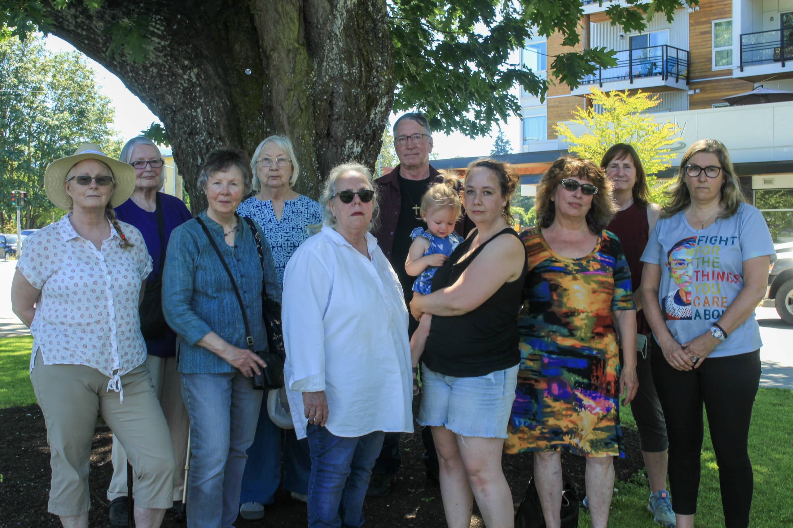 A group of women (and one man) stand in front of a tree in Downtown Duncan posing for the photo. One of the women is holding her baby. Everyone in the photo has a serious expression on their face. They represent a group of abortion advocates concerned about the opening of House of Grace in the community.