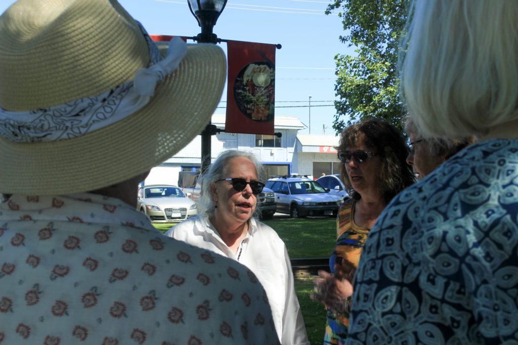 The foreground of the photo is taken up by the backs of two women. Half of their body is in frame and the other half is off-frame. The photo shows their backs from shoulder up. One of them is wearing a sunhat. In between the two, in the background of the photo, is a clear image of Rhoda Taylor wearing a white shirt and speaking to the women. Next to her on the right is Gayle Hurmuses.