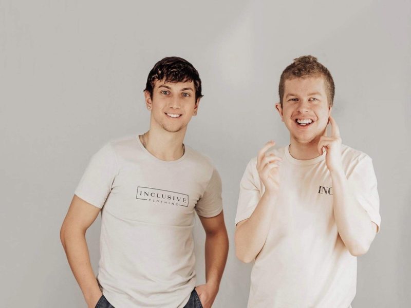 VIU student Brant Cartwright, left, stands beside his brother Riley with a white backdrop smiling in Inclusive Clothing T shirts