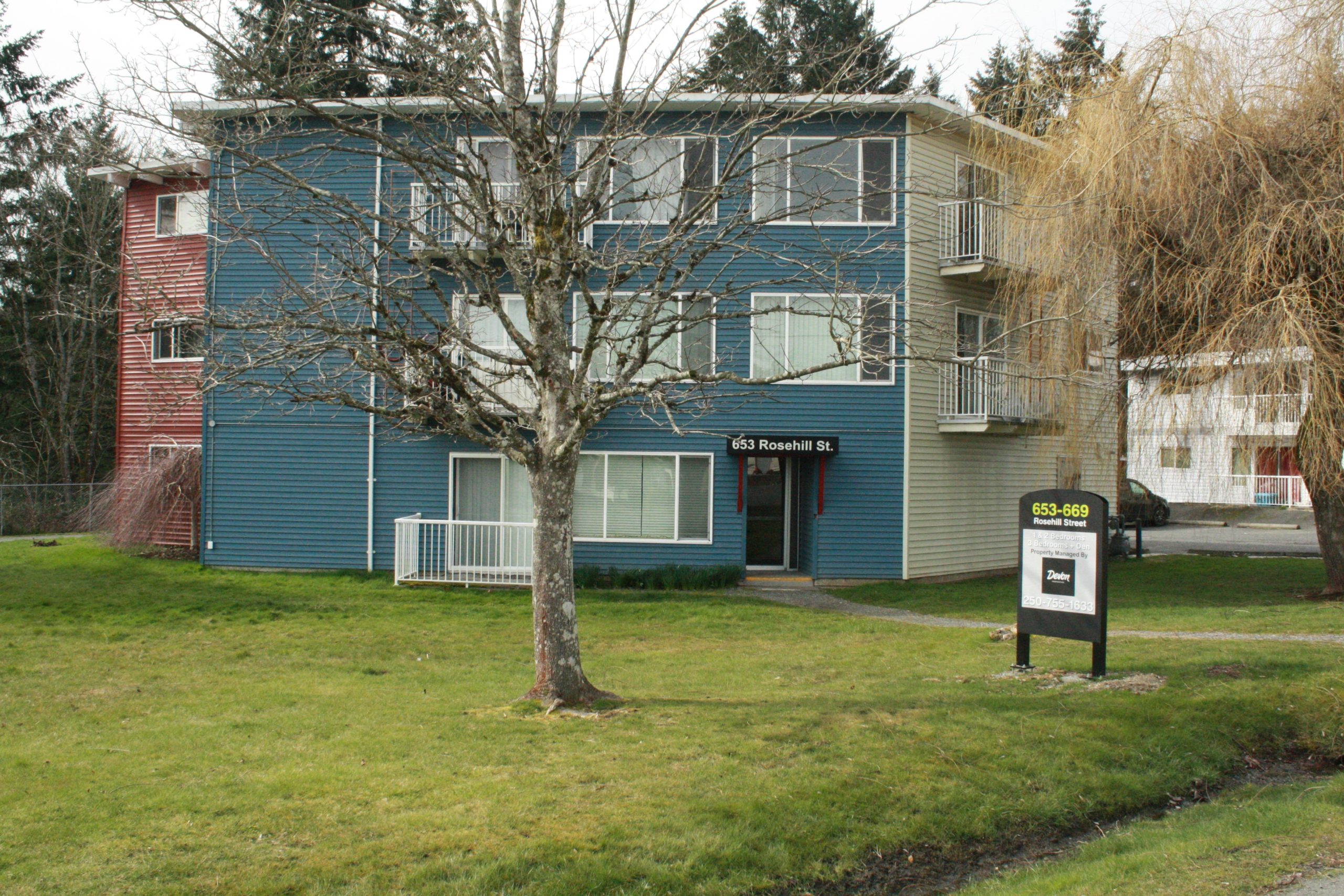 A blue apartment building with a green lawn displays a vacancy sign in front marked Devon Properties.