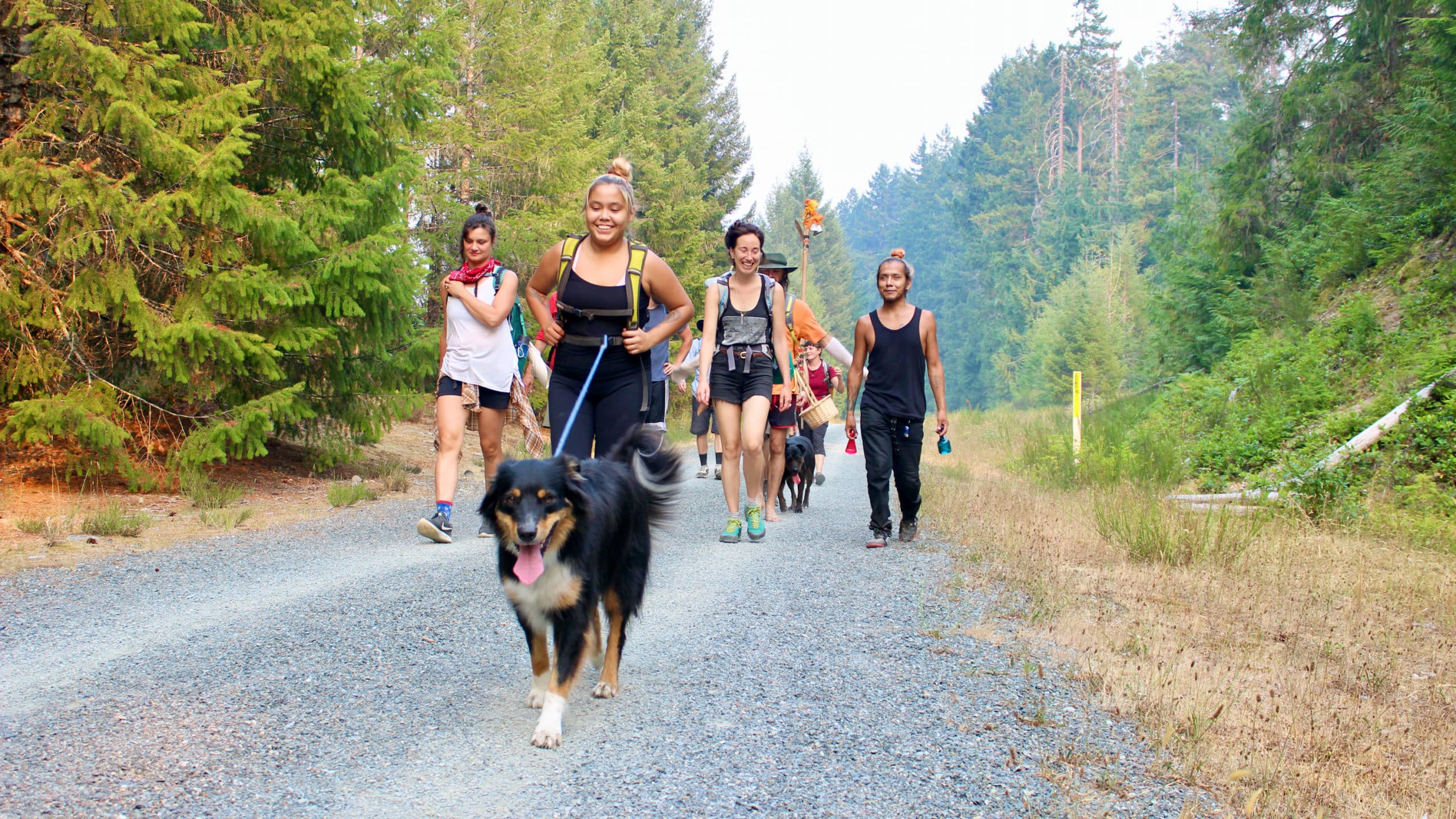 Smiling people walk down a wide trail with a happy dog in front.