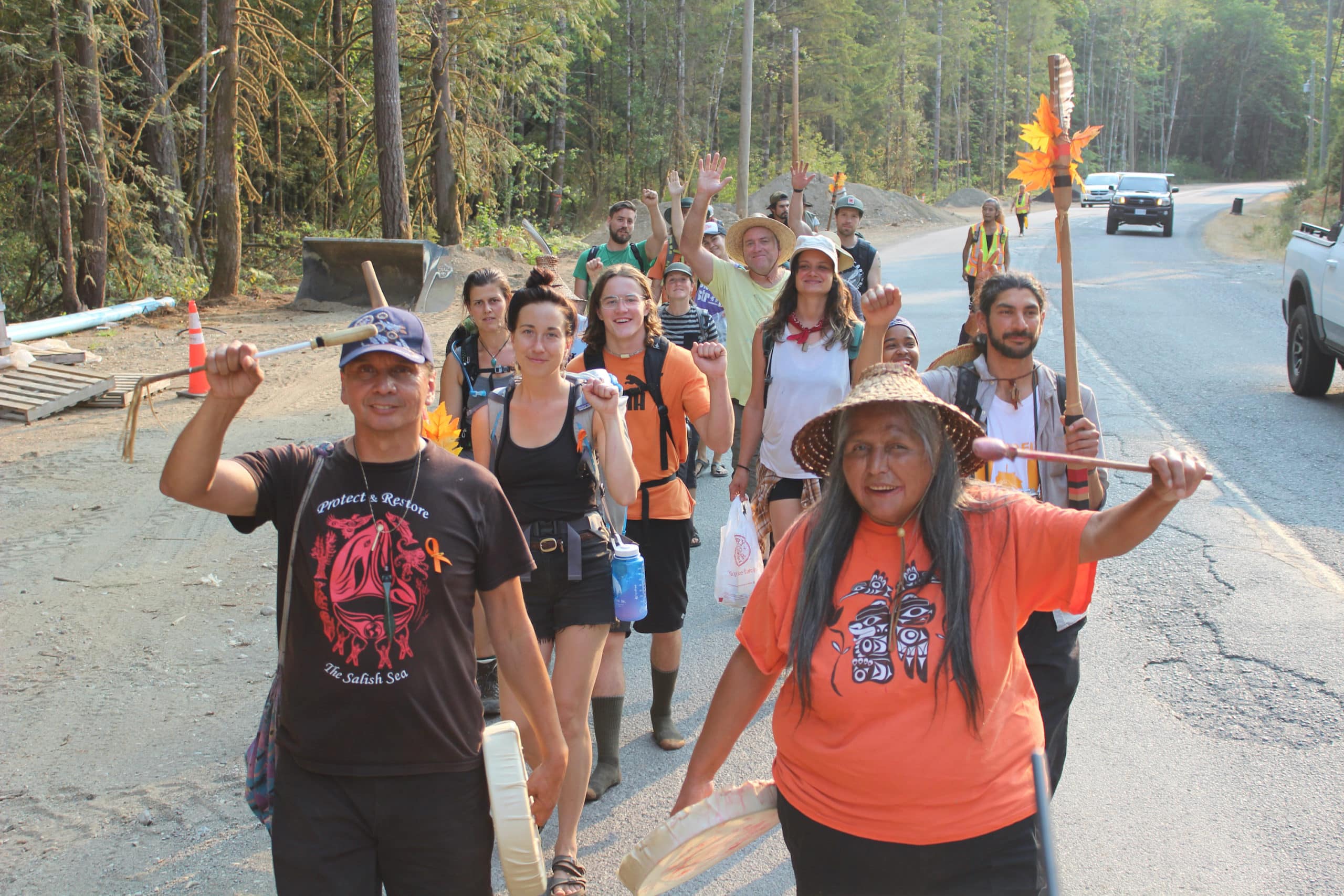 A group of about 20 people walk on the shoulder of a road. Many hold a raised fist. 