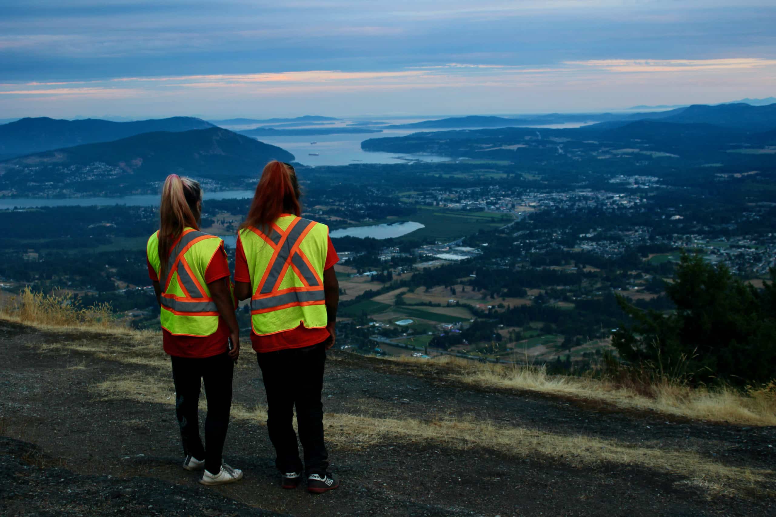 Seen from behind, two people in high-visibility vests look out from a mountain top.