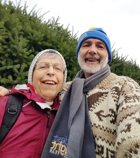 A woman and man embrace with winter hats and scarves on next to a hedge.