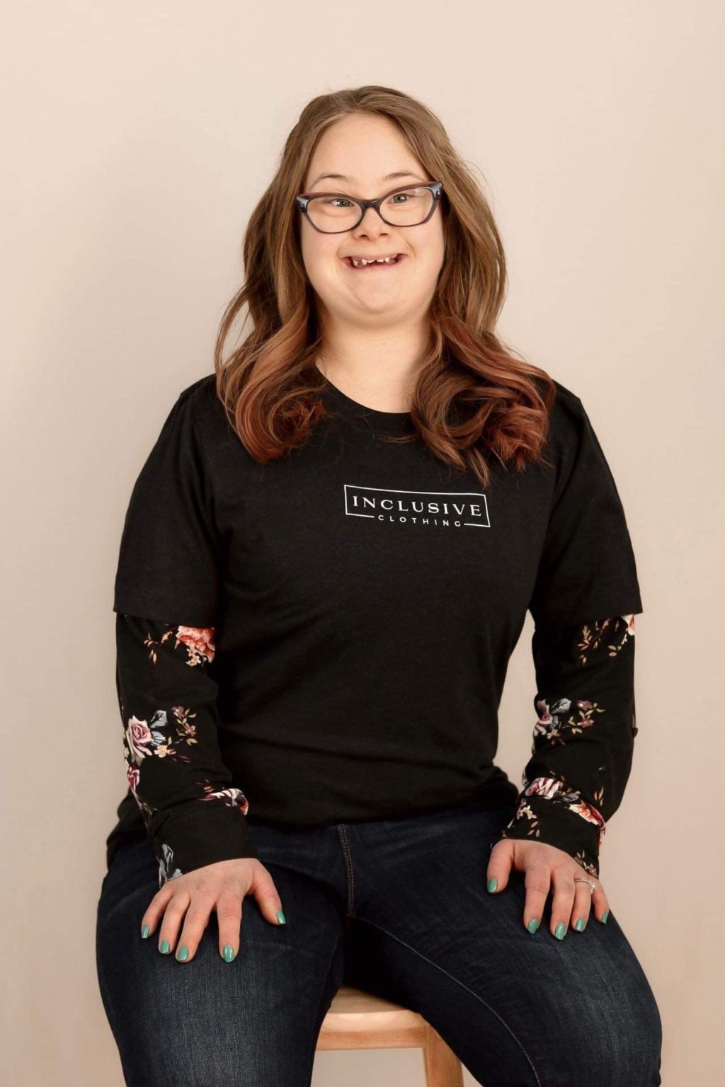 Mikaela models a black Inclusive Clothing T-shirt sitting on a stool with a white backdrop. She's a lifelong friend of  Brant from his hometown community, she has beautifully curled red hair a big smile.