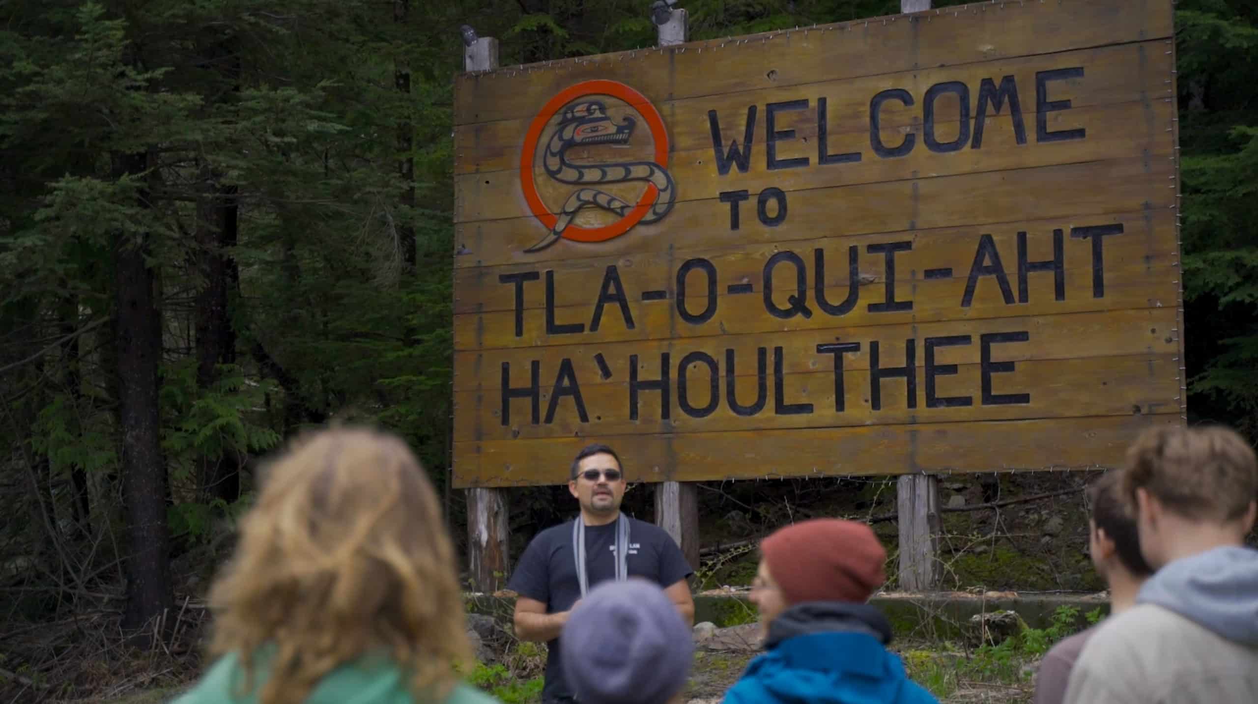 Eli Enns stands in front of a Welcome to Tla-o-qui-aht Ha'Houlthee sign and welcomes a group of students.