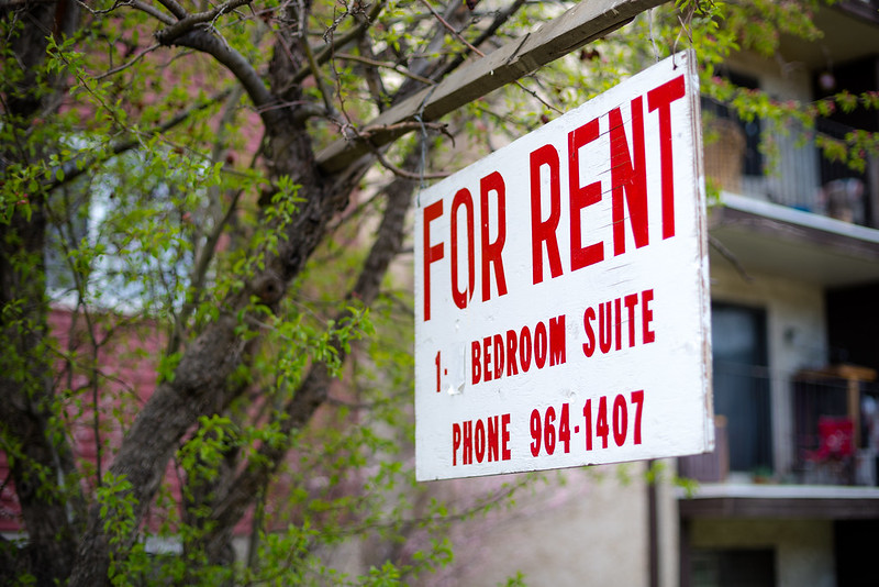A sign reads For Rent in front of a building with a tree beside it.