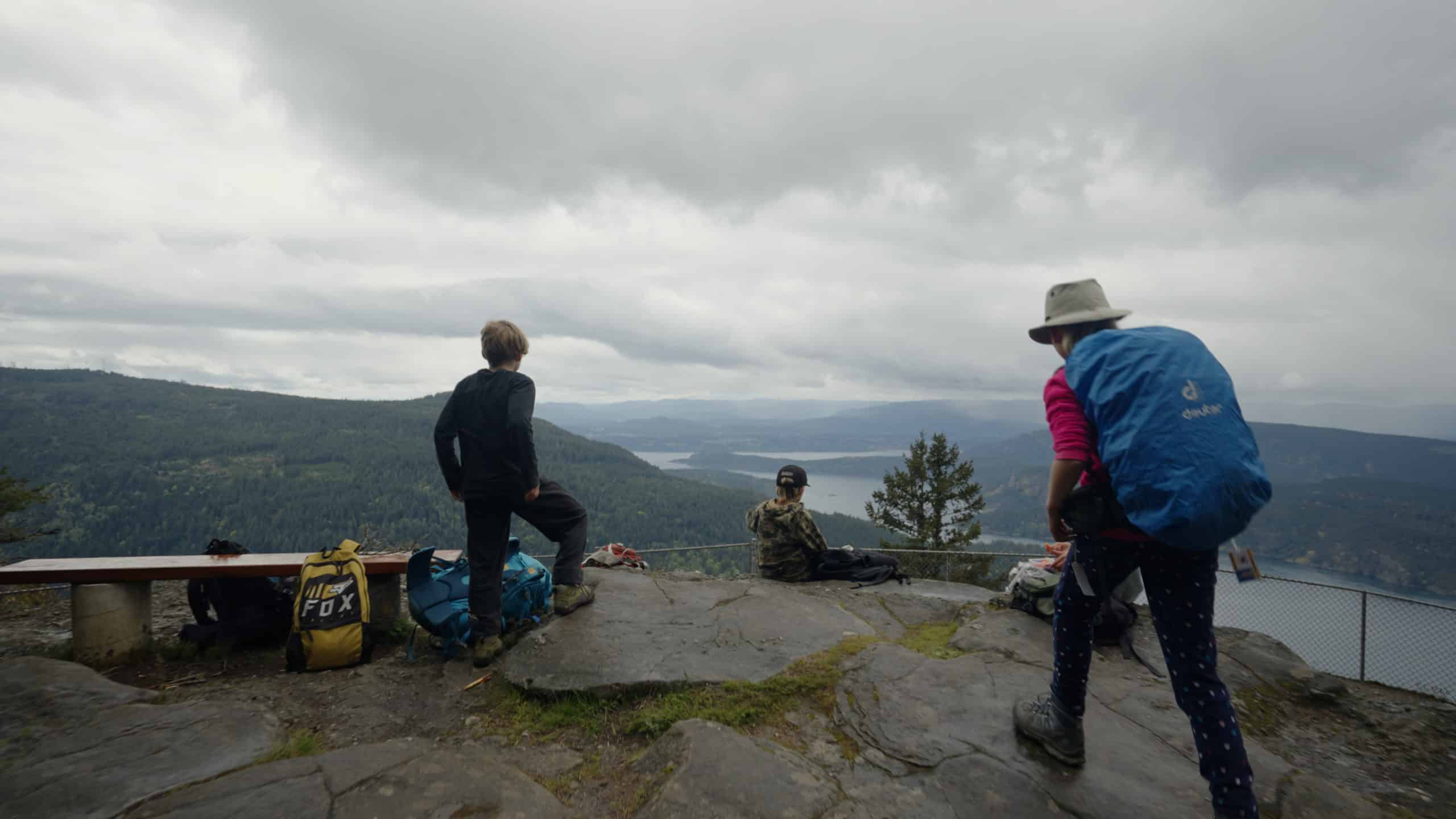 The photo captures the backs of two children and one adult-looking figure on top of a mountain overlooking the Cowichan Valley. The child on the far left is standing. A child further into the background of the photo is sitting in the middle and the figure on the right is walking towards the children.