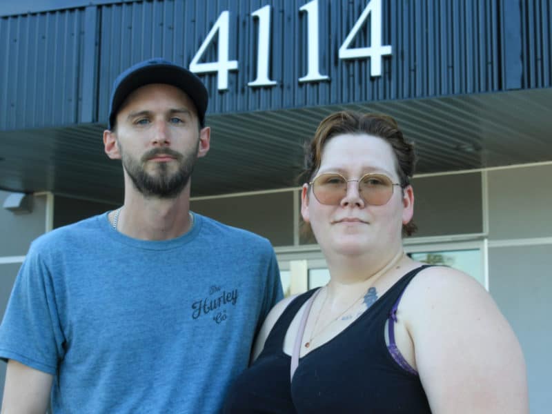 A man and a woman stand, looking serious, in front of a building facade.