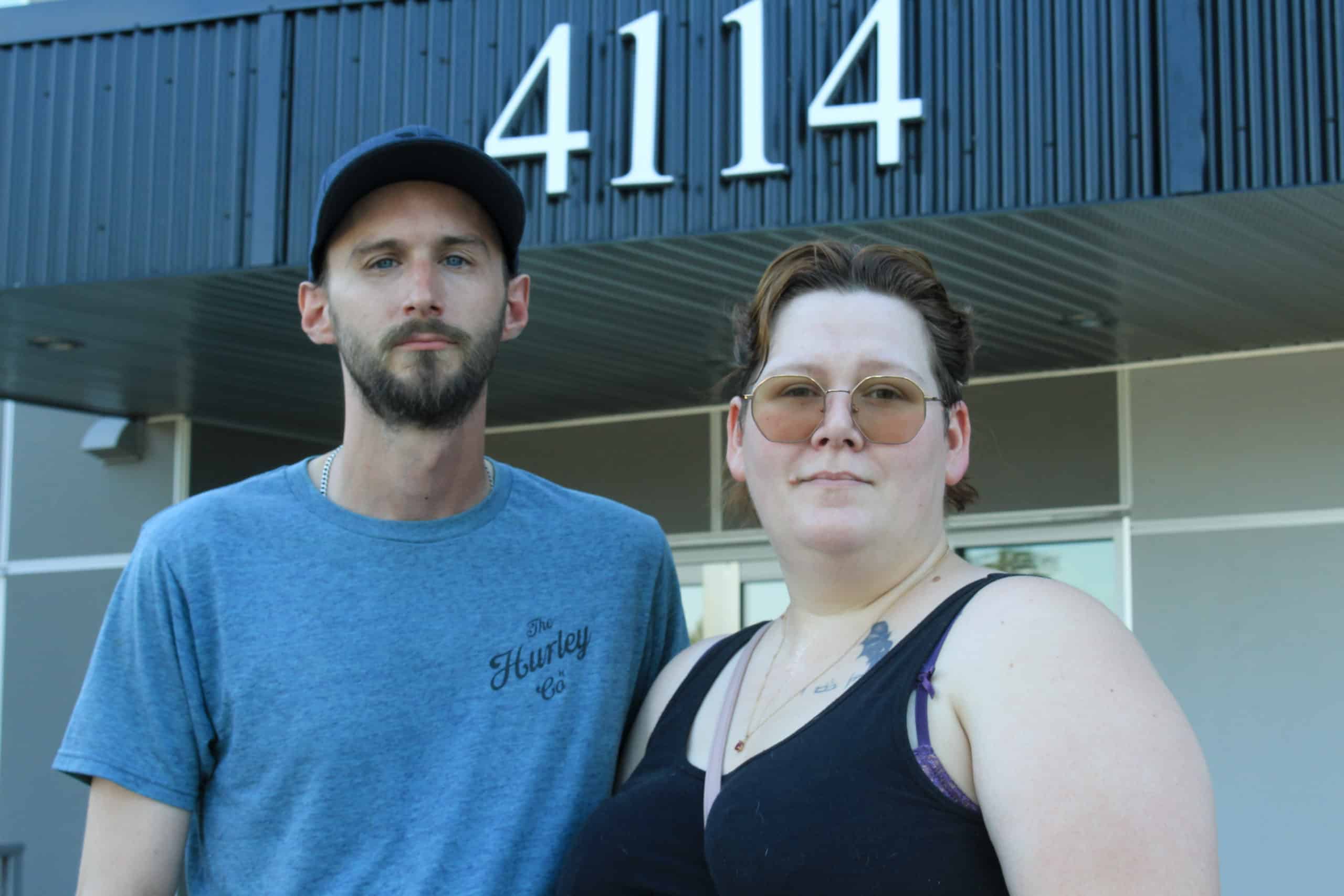 A man and a woman stand, looking serious, in front of a building facade.