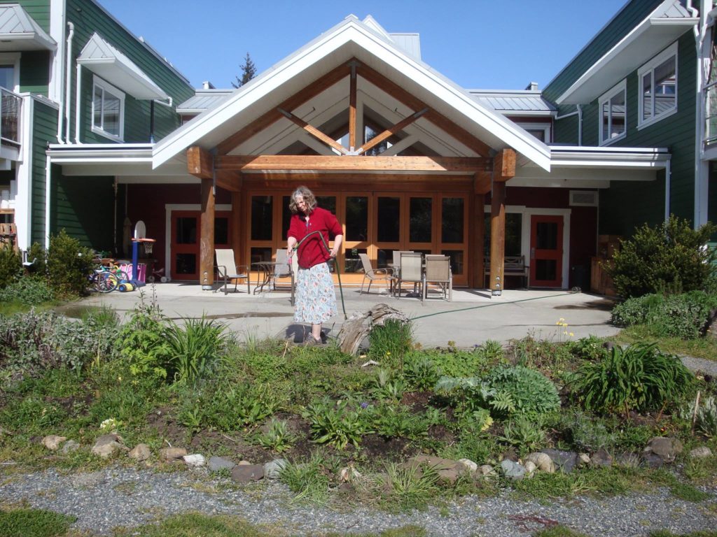 Pacific Gardens Cohousing features an outdoor common space for residents to gather. A person is shown watering the garden in front of a community dining patio.