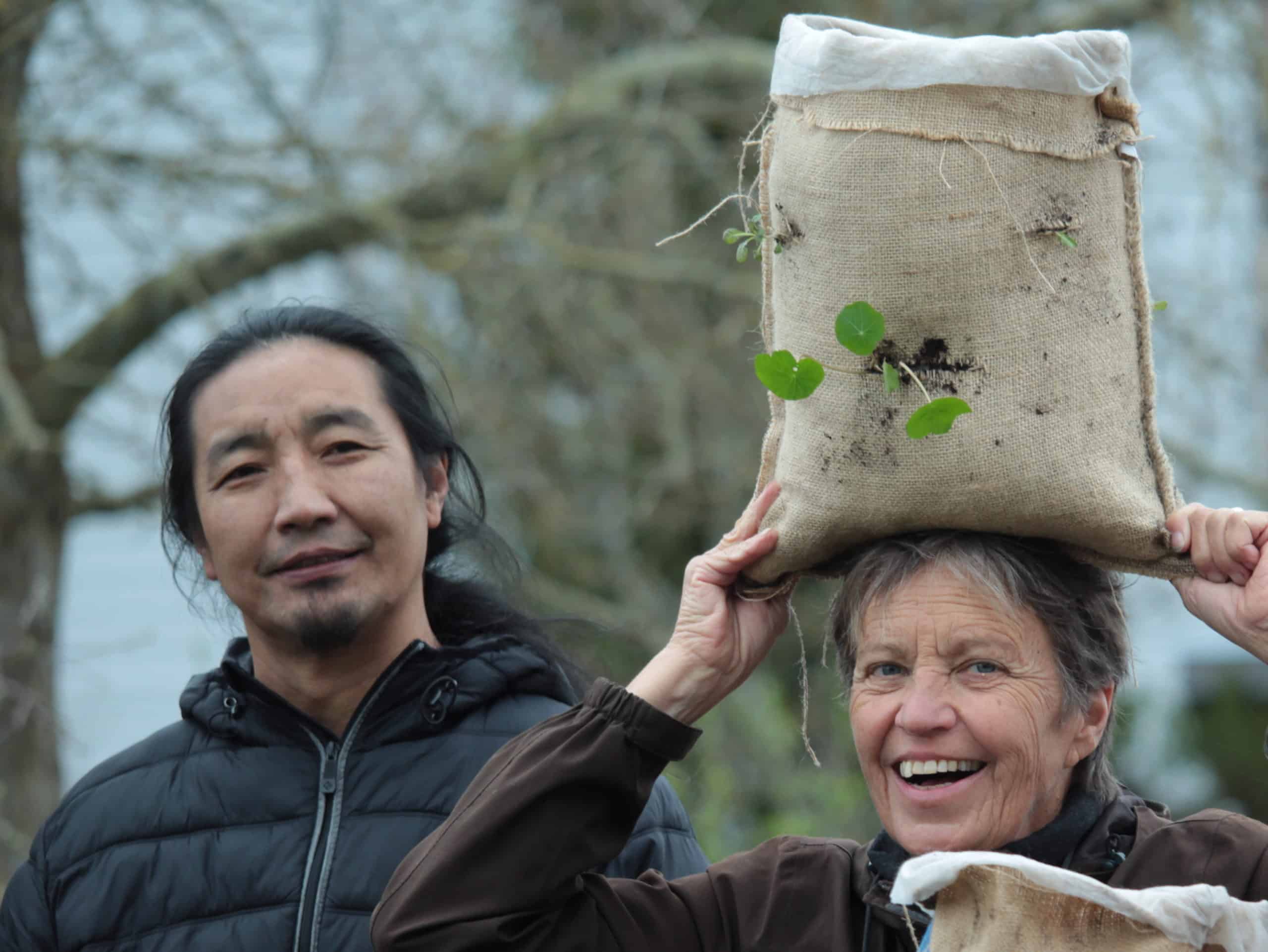 Pema Rigzin and Elke Cole smile as Elke holds a mini burlap sack garden on top of her head.