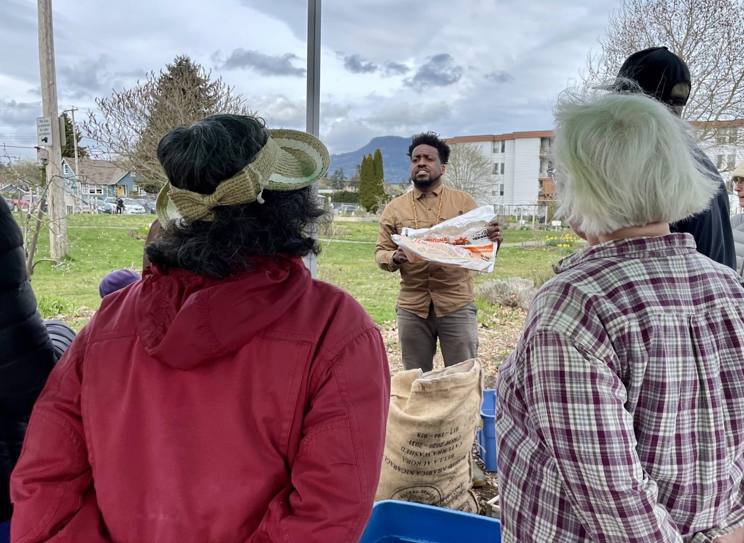 Ariel Reyes Antuan of IYÉ Creative leads a demonstration on how to build and install burlap sack gardens in the background. In the foreground are the backs of community members who are watching Ariel.