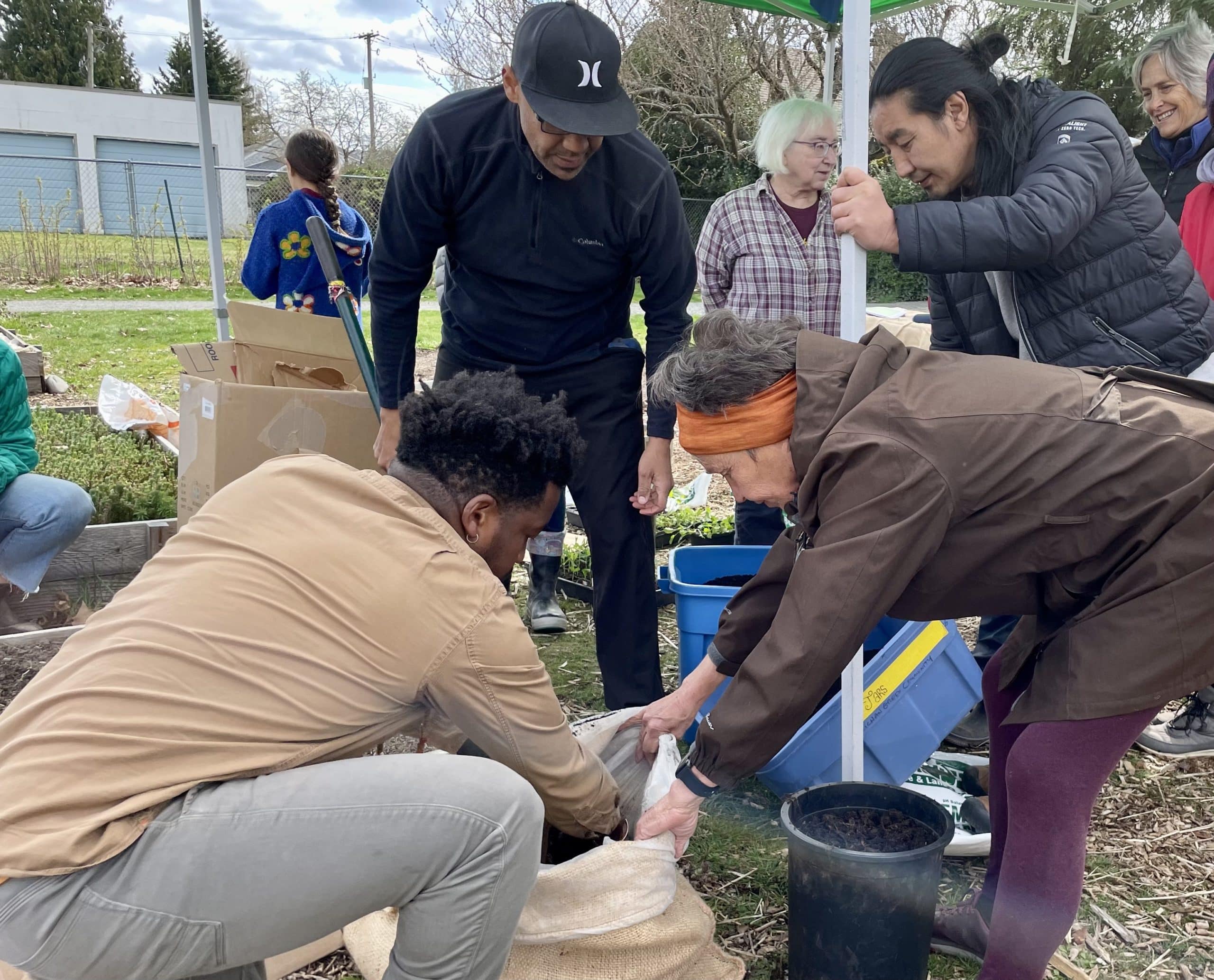 Community members crouch, kneel and stand as they fill a burlap sack with soil.