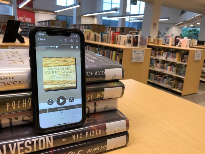 A stack of books sits on the table with a phone showing an audiobook on the screen.