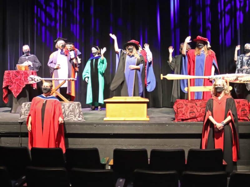 People in academic robes and face masks stand on stage for a COVID-style grads ceremony at VIU 2021.