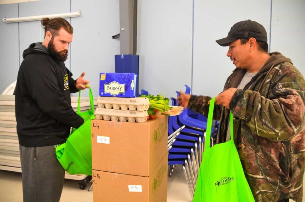 Two men stand in front of a table and fill shopping bags with food items.