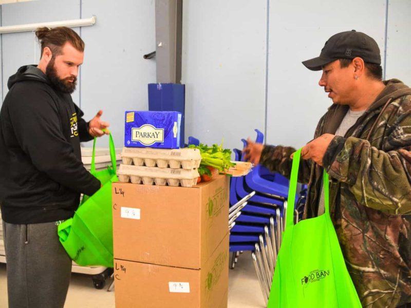 Two men stand in front of a table and fill shopping bags with food items.