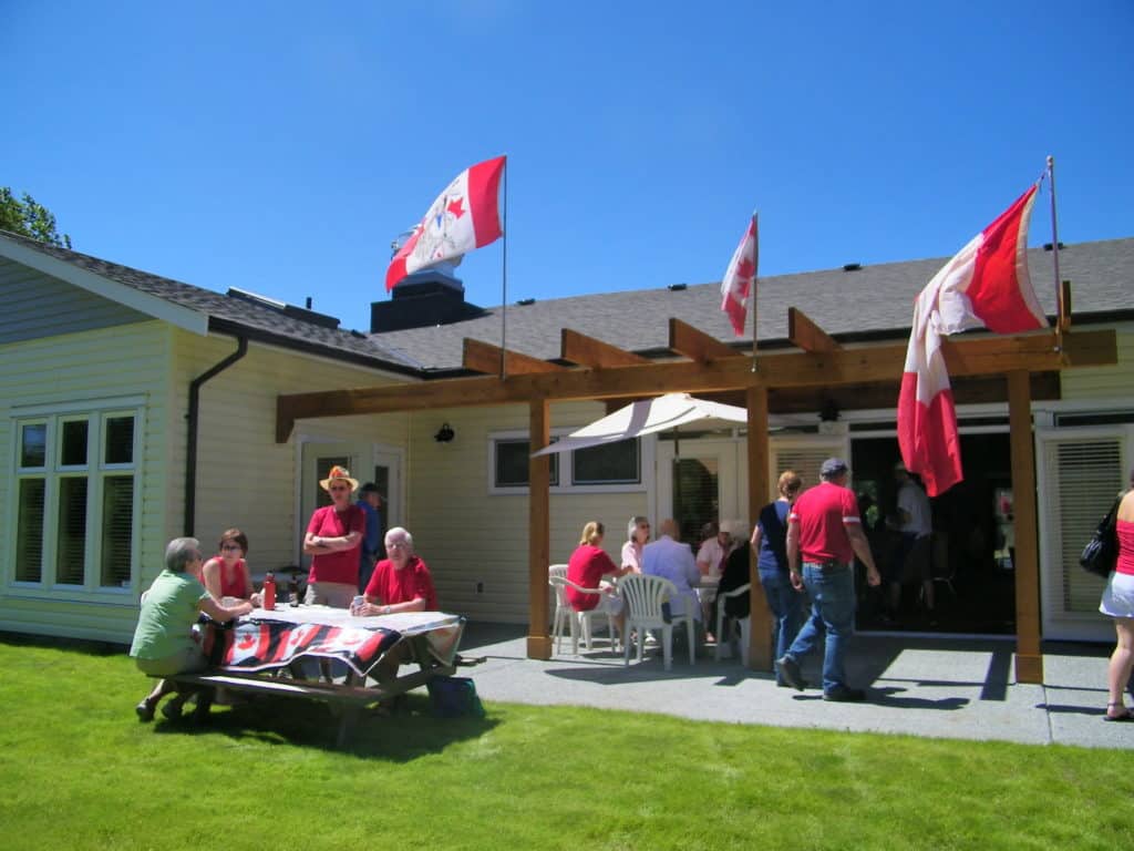 A community hall is shown with Canada flags and seniors sitting at picnic tables in front on the lawn.