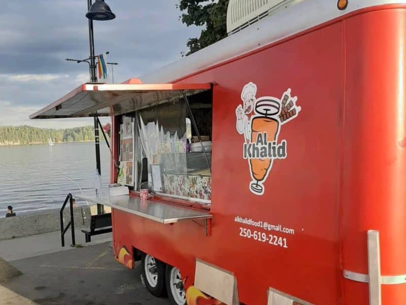 A red Shawarma food truck with the words Al Khalid is parked at a beach.