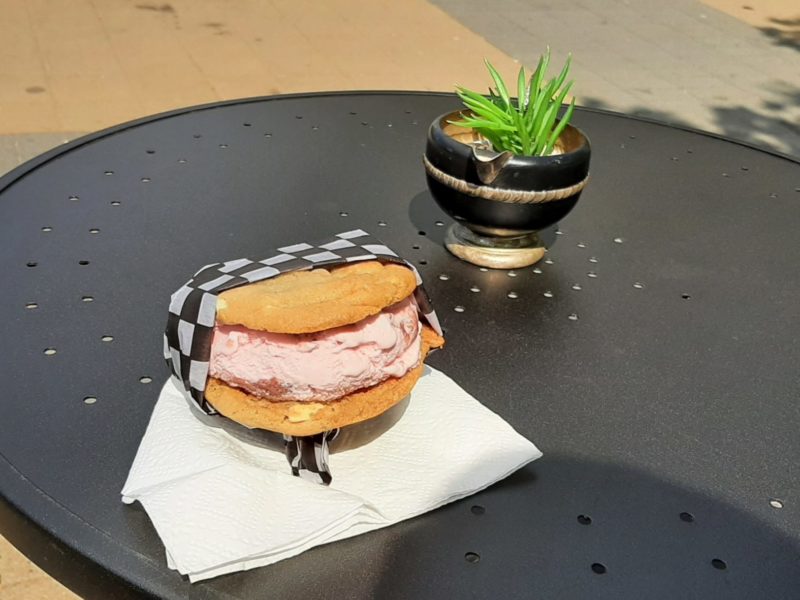 An ice cream sandwich, oatmeal cookie layers with pink ice cream in between, sits wrapped in black checkered wax paper on a black bistro table.