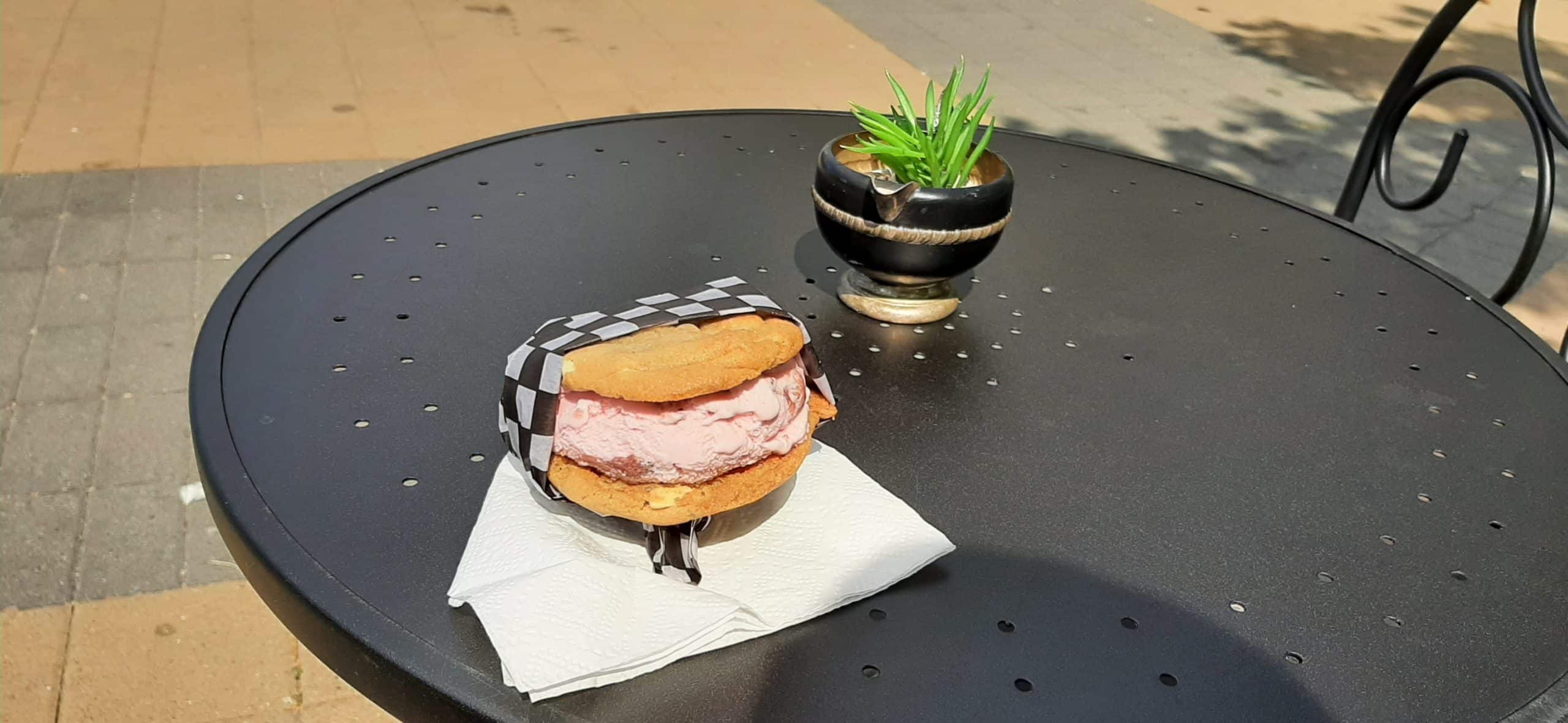 An ice cream sandwich, oatmeal cookie layers with pink ice cream in between, sits wrapped in black checkered wax paper on a black bistro table.