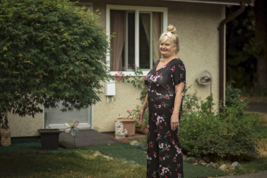 A senior stands in front of her unit with a house dress and a smile.