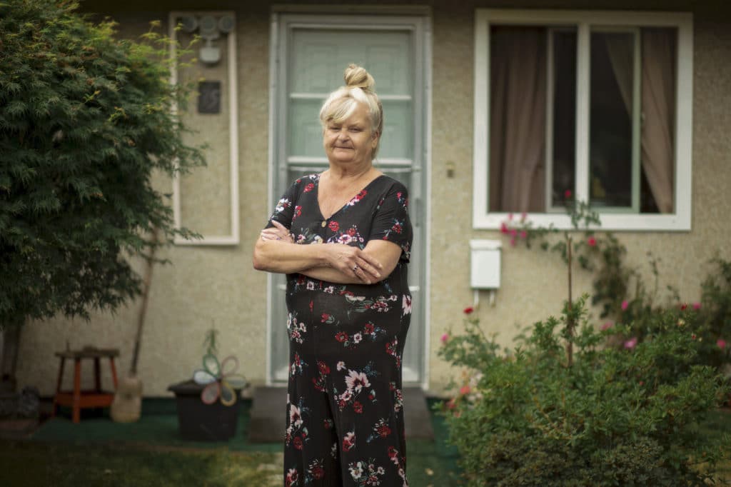 A blonde person with a bun stands in front of a front door on the lawn with arms crossed.