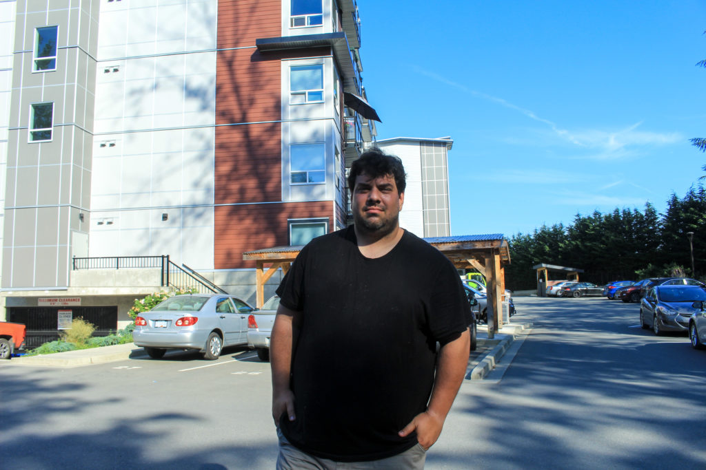 John Voutier is pictured from the waist up and standing in the parking lot behind the Magdalena building. Behind him on the left is the corner of the building and some vehicles parked in front of it. On the right is blue sky. John is wearing a black t-shirt.
