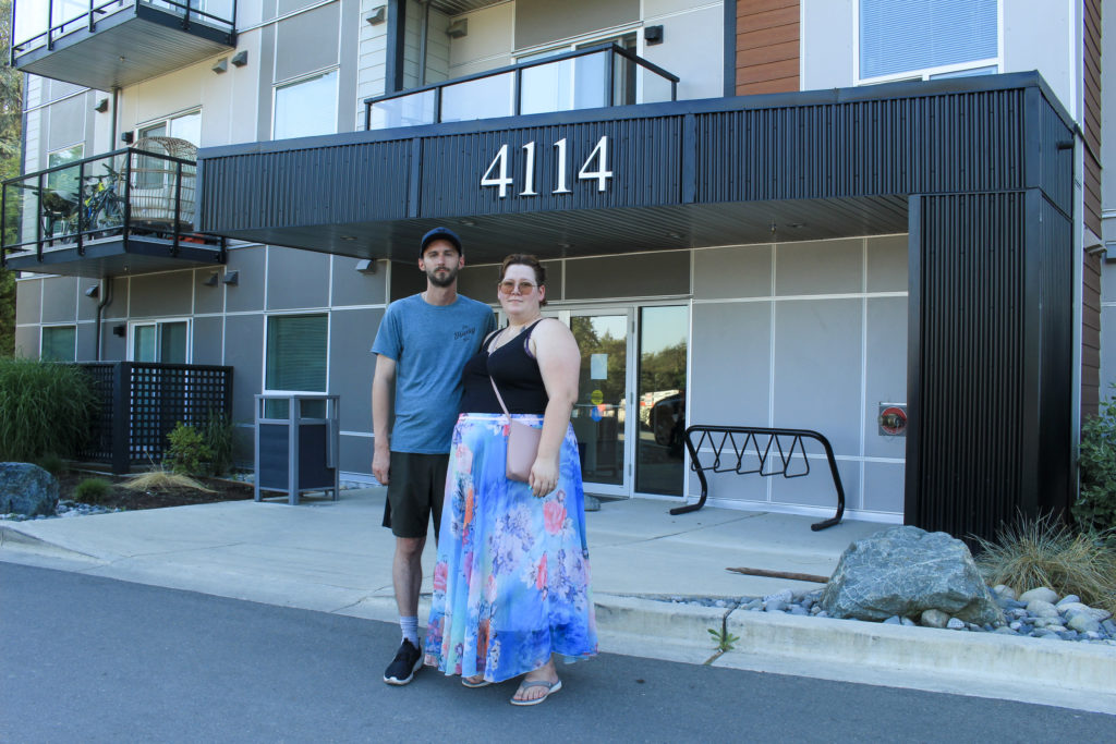 Sean and Jess Phair stand side-by-side with one arm around each other. They are standing on pavement and behind them is the entrance to the Magdalena building. Above their heads and behind them is the building's address marker, with large white numbers that say 4114. Sean is wearing a blue t-shirt and black shorts. Jess is wearing a black tank top and long blue and orange skirt.