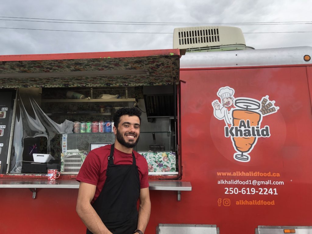 A person in an apron smiles in front of a bright red food truck with the word Al Khalid