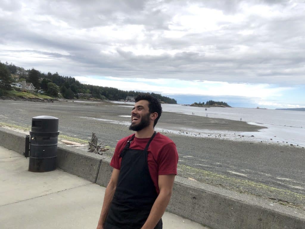 A man with short hair and a beard smiles on a walkway at the beach, wearing a black apron and red shirt.