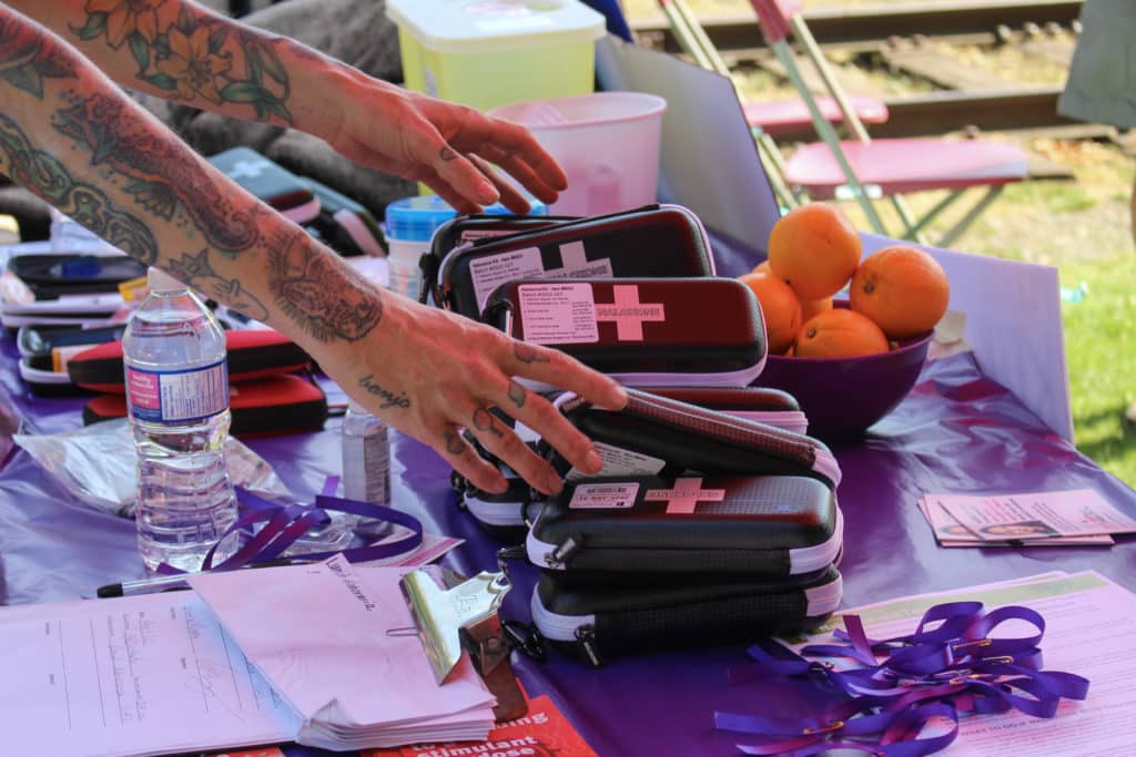 Tattooed arms reach towards a pile of naloxone kits on a table.