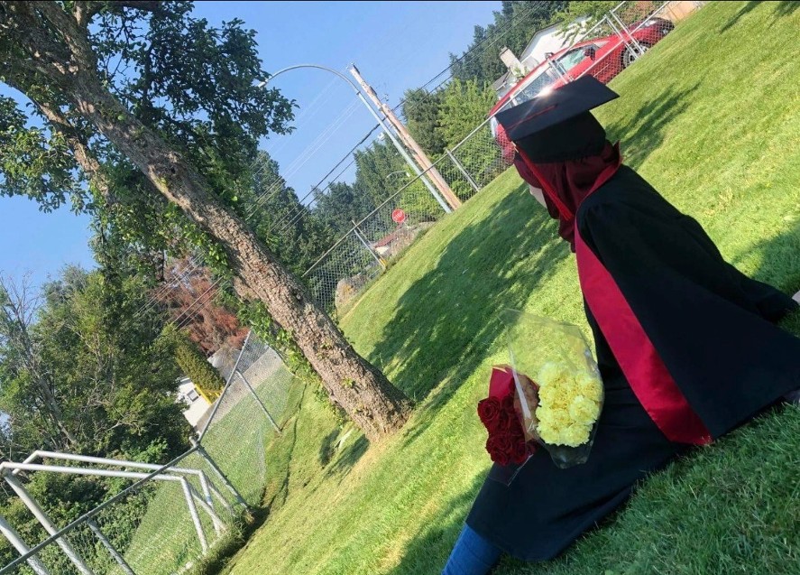 A person in black and red graduation robes sits in the grass with a tree in the foreground.