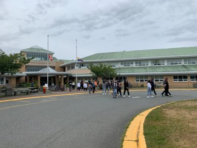 A line of students walk out of the front entrance to a school building and across a street. All seem to be ages 14-18, some are wearing masks.