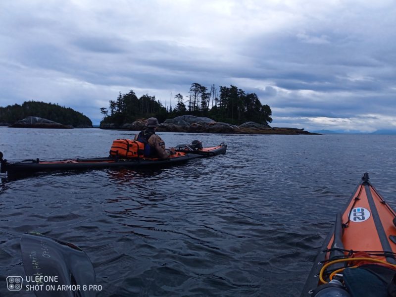 The tip of a kayak is shown with a full kayak to the left of frame on the ocean with a forested island in background.