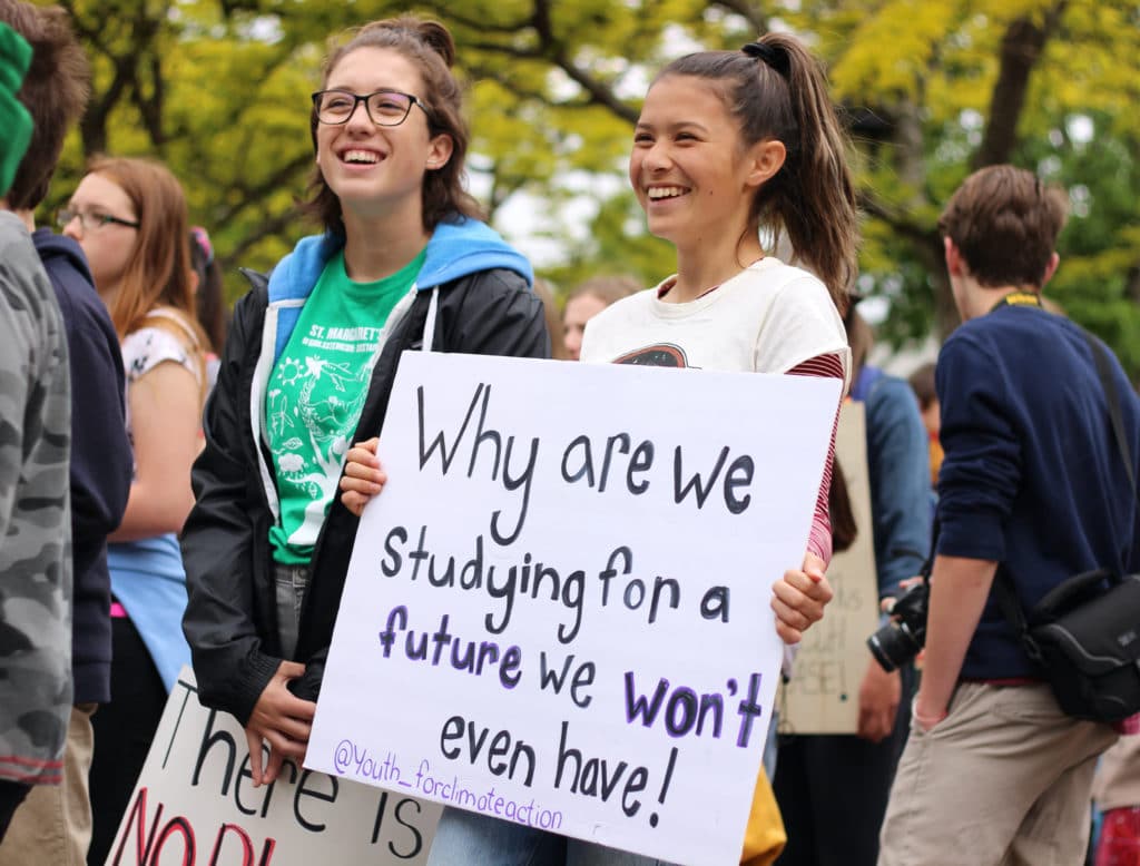 Two young people stand in a crowd holding hand-drawn signs. One sign reads, “Why are we studying for a future we won’t even have!”