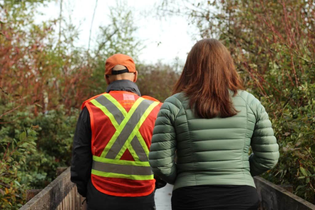 The backs of Paul Gowland and Phaedra Douglass are seen, walking on a boardwalk surrounded by forest. Paul is slightly ahead of Phaedra.